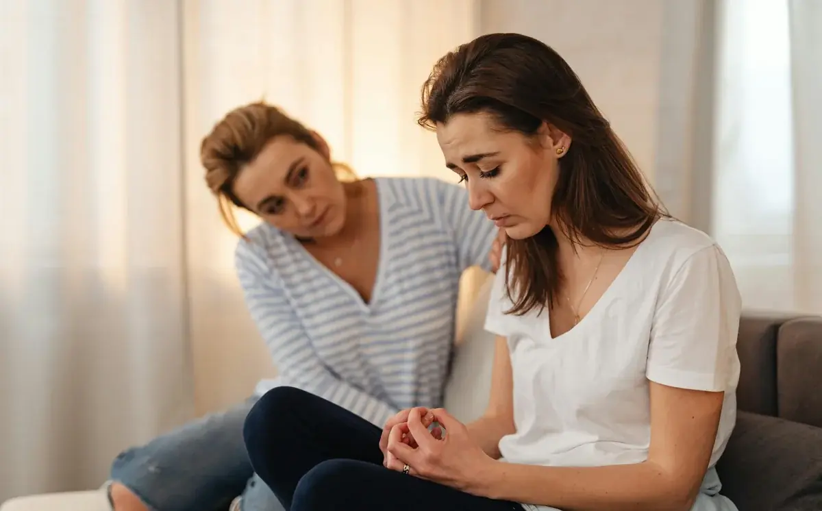 A friend sits beside a grieving woman on a couch, hand resting gently on her shoulder. The grieving woman is looking down with hands clasped in her lap, soft daylight from a window behind them. The kind of quiet showing-up the Supporter's Guide teaches.