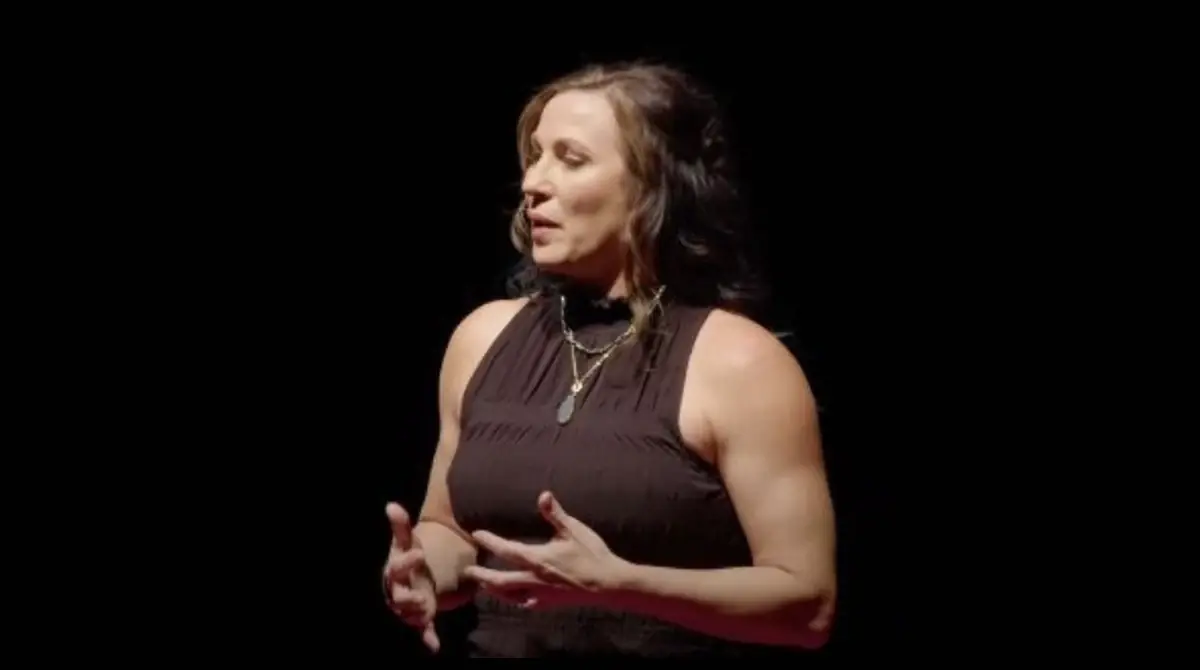 A woman speaking onstage in warm theatrical lighting, gesturing with both hands as she shapes a thought aloud. The voice work behind the Three Guided Audio Meditations.